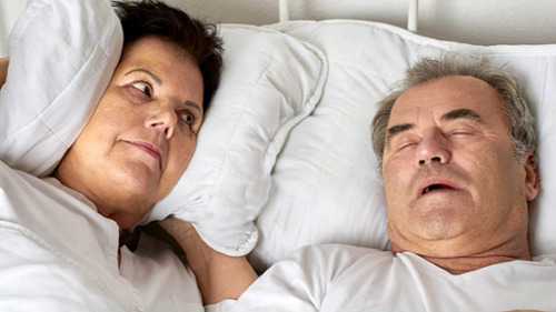 Woman covering ears with pillow while partner snores loudly in bed at night